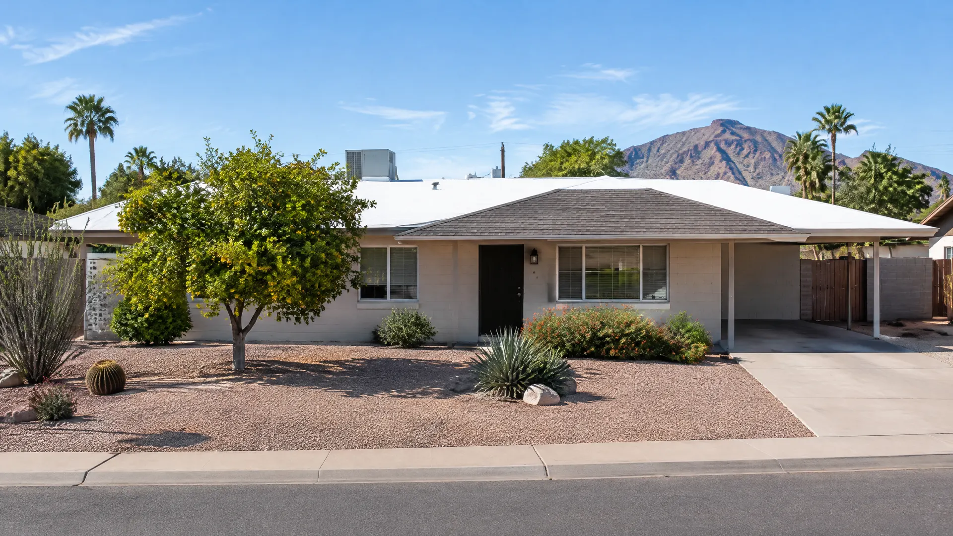 Modest 1960s single-story block ranch home in west Tempe, Arizona with a low-slope roof partly flat with a white foam coating and a shingled front section, desert landscaping and a citrus tree