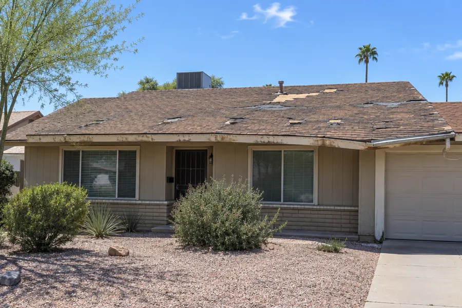 Single-story Tempe ranch house with an old worn roof before replacement: faded curled brown asphalt shingles with bare patches, stained fascia, sagging gutter, sunny day, front view