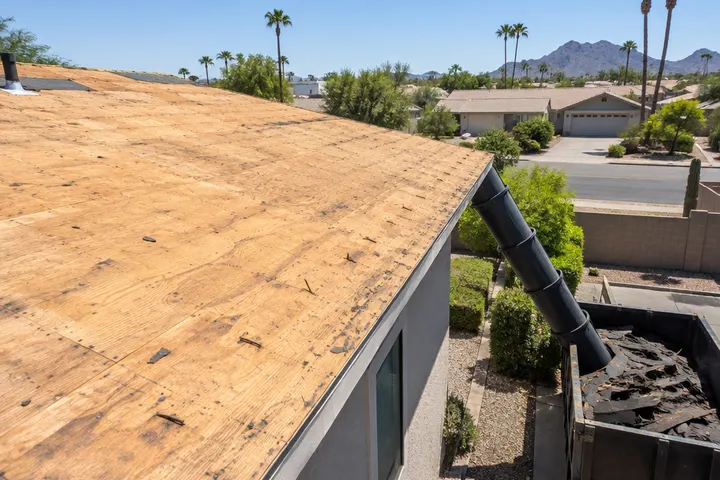 Roof deck stripped of old shingles, bare plywood and a few old nails, a debris chute leading to a dumpster below, no faces